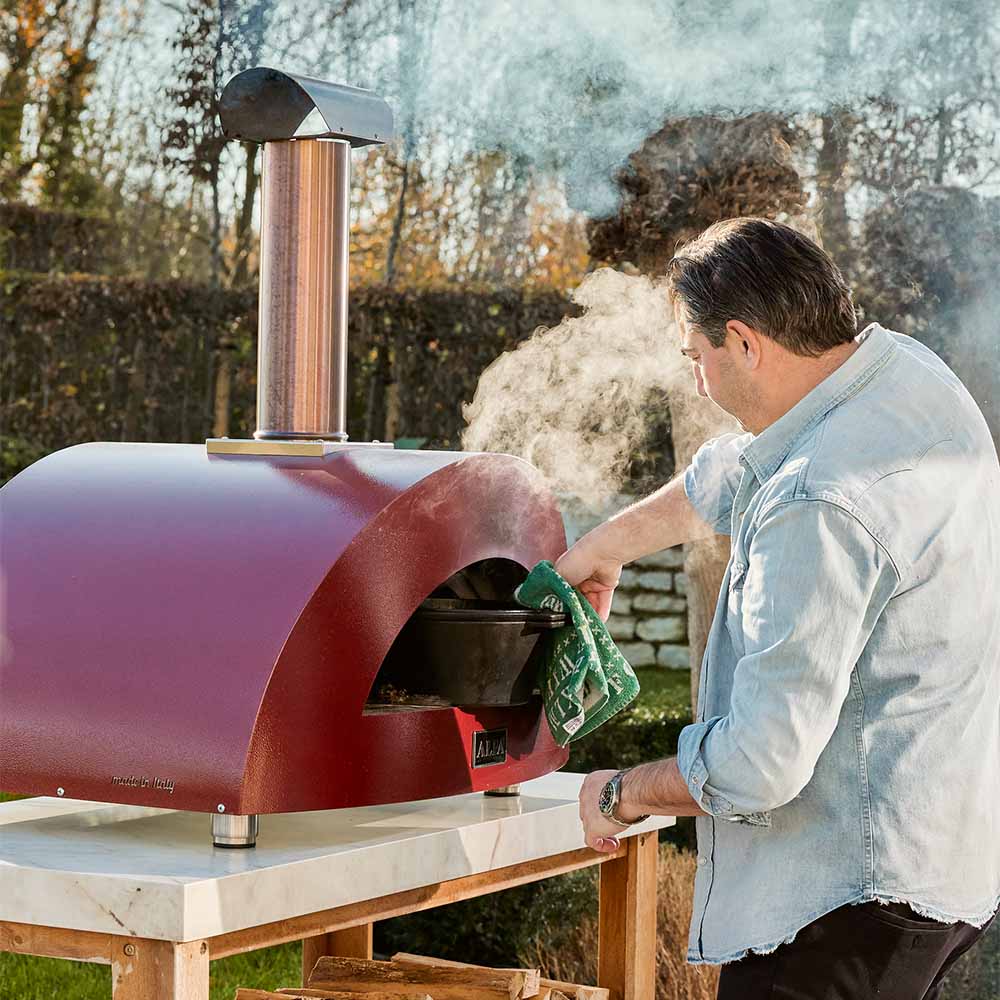 Red wood-fired pizza oven on a stand with smoke rising from the open arch