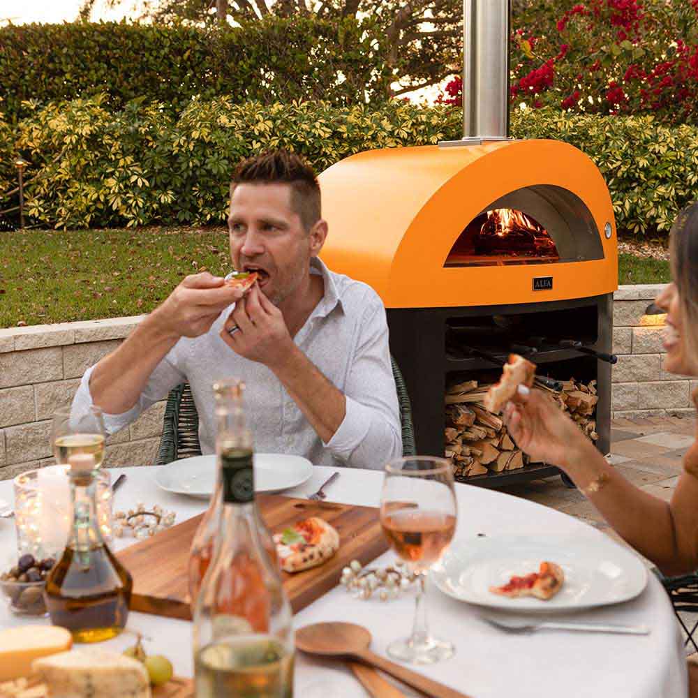 Freestanding gas pizza oven with yellow hood, burning firebed, and pizza near the opening