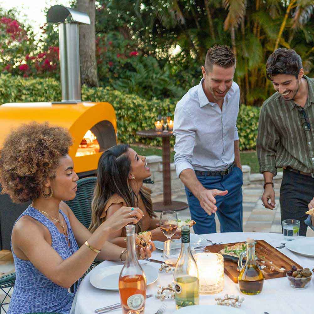 Group meal at a yellow gas pizza oven; chimney and open oven mouth visible.