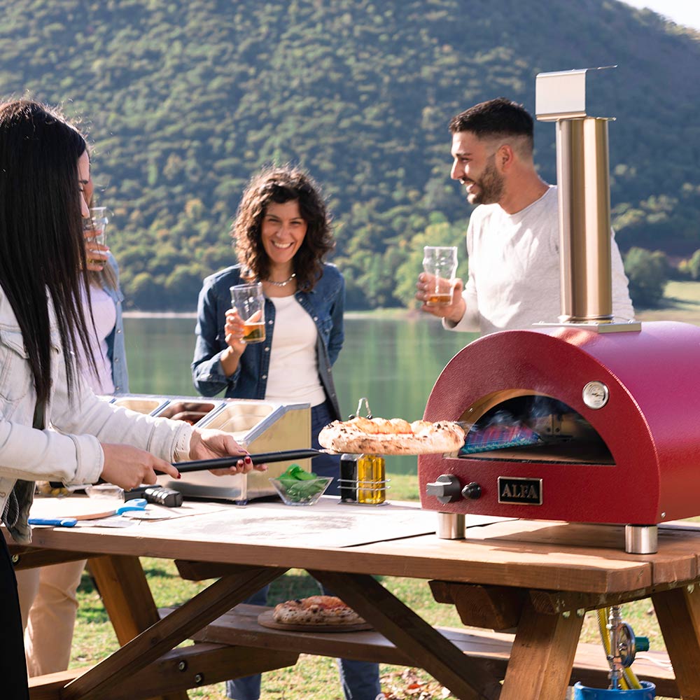 Portable gas pizza oven with red dome and tall chimney on an outdoor table