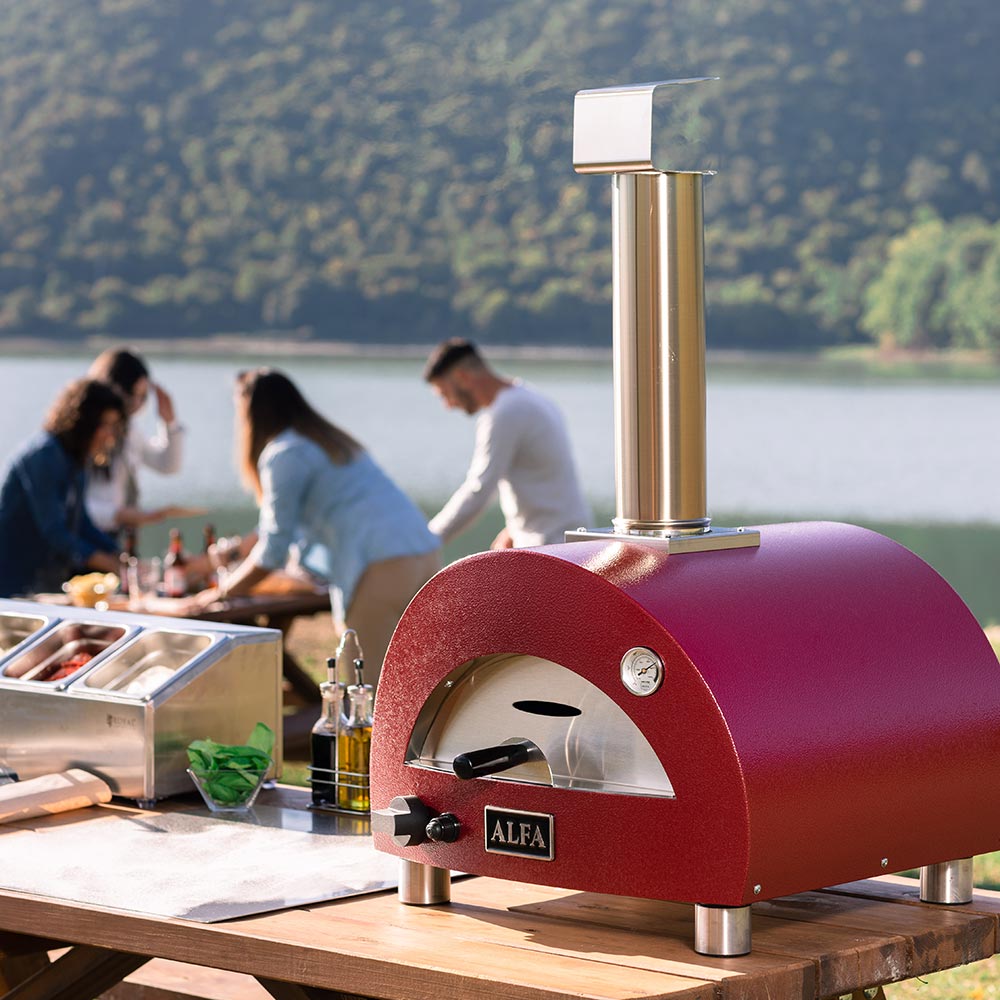Portable gas pizza oven with red dome and tall chimney on a wooden outdoor table