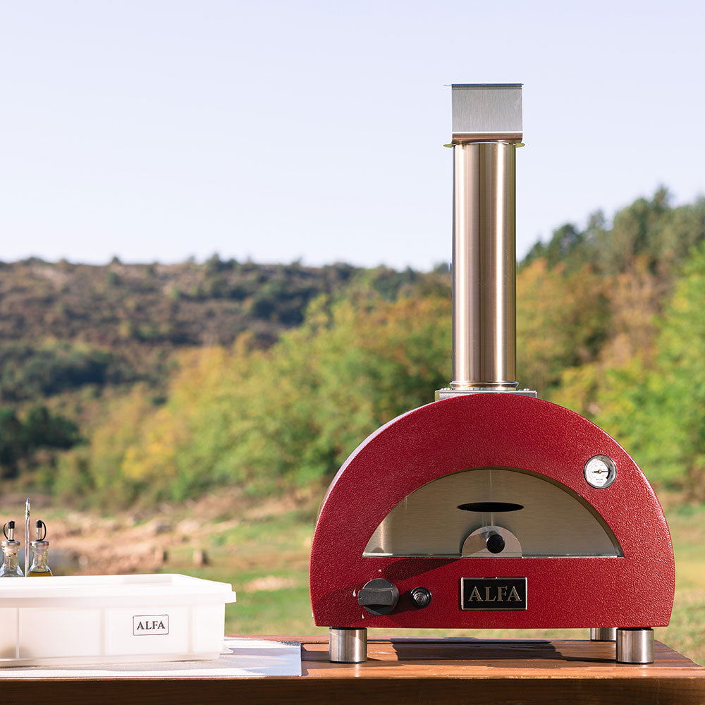 Portable gas pizza oven with red dome, gray firebox opening, and tall chimney outdoors