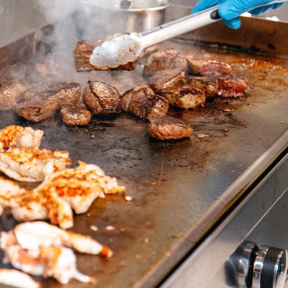 Stainless griddle cooking surface with browned meat pieces and blue-gloved tongs