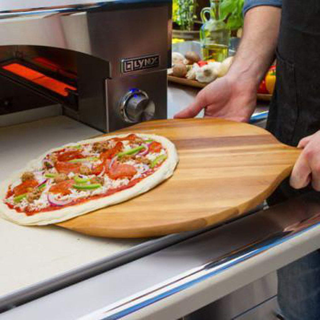 Hand loading pizza onto a wooden peel into the open Lynx pizza oven