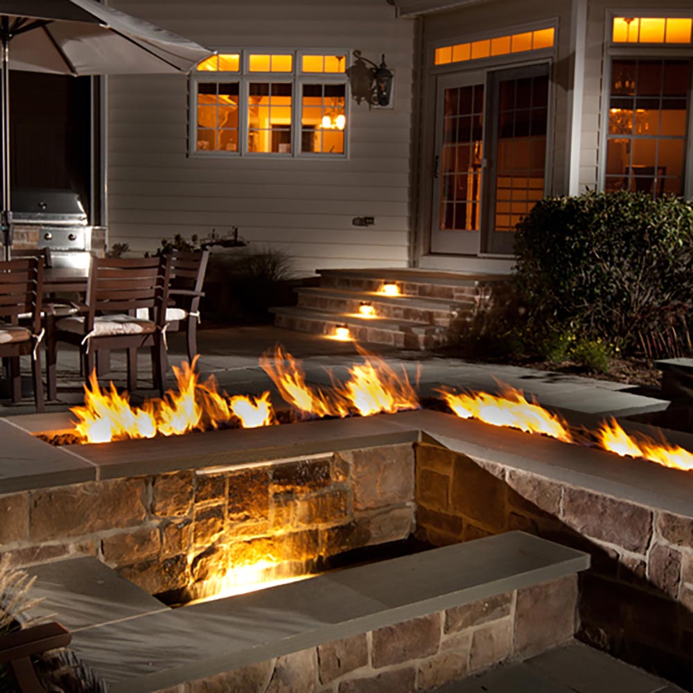 Long linear fire pit burner with flames running across the raised stone surround