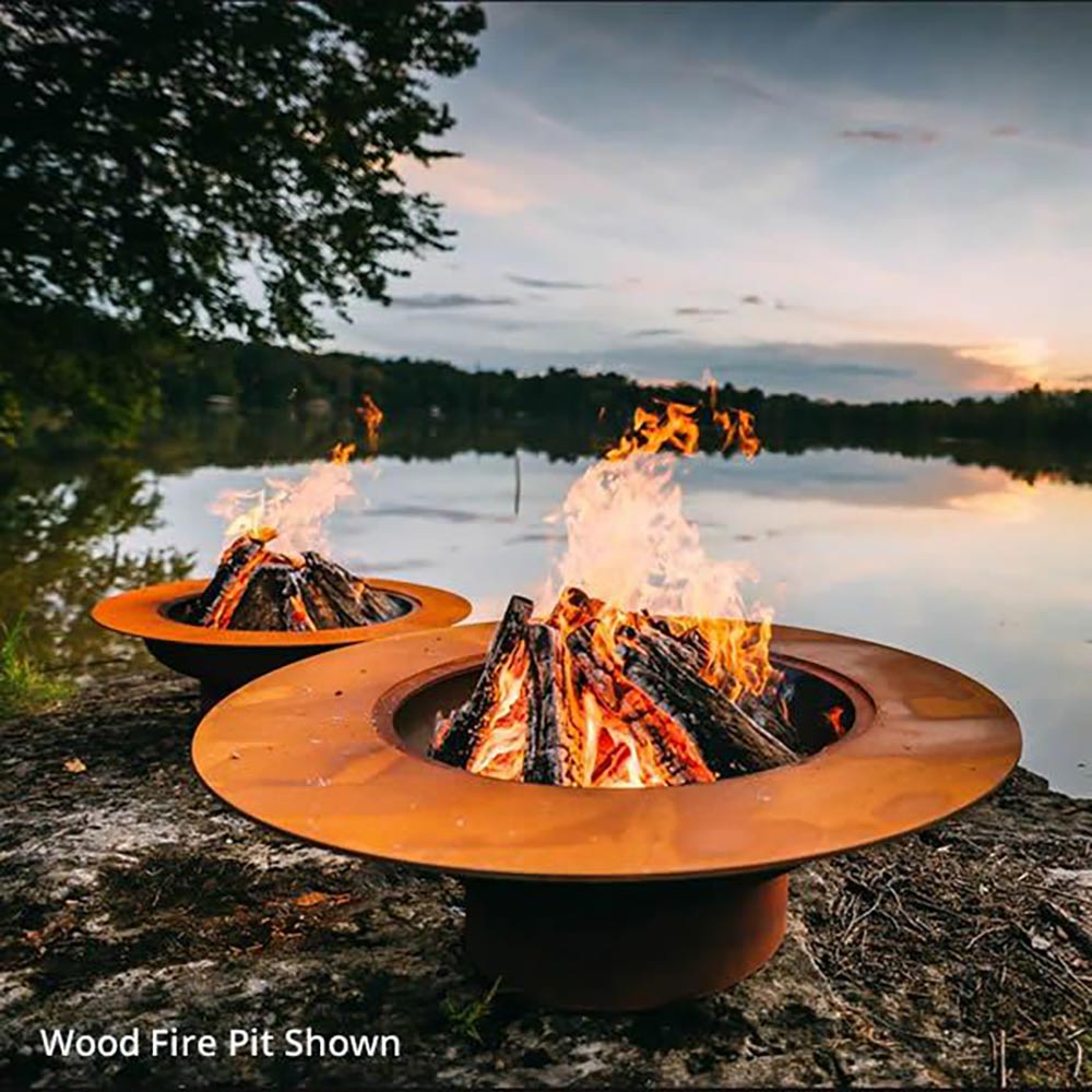 Round gas fire pit with flames rising from central burner, near a lake