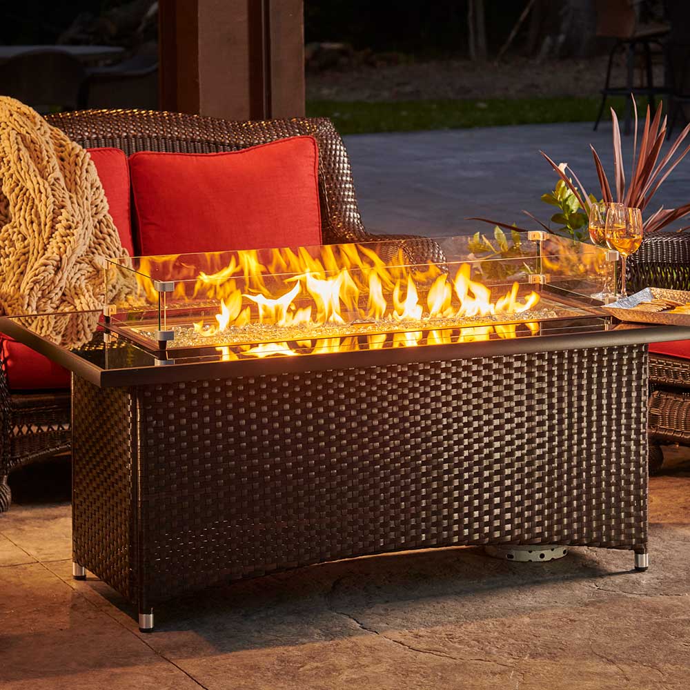 Outdoor gas fire table with glass windscreen, visible flames, and woven dark brown sides