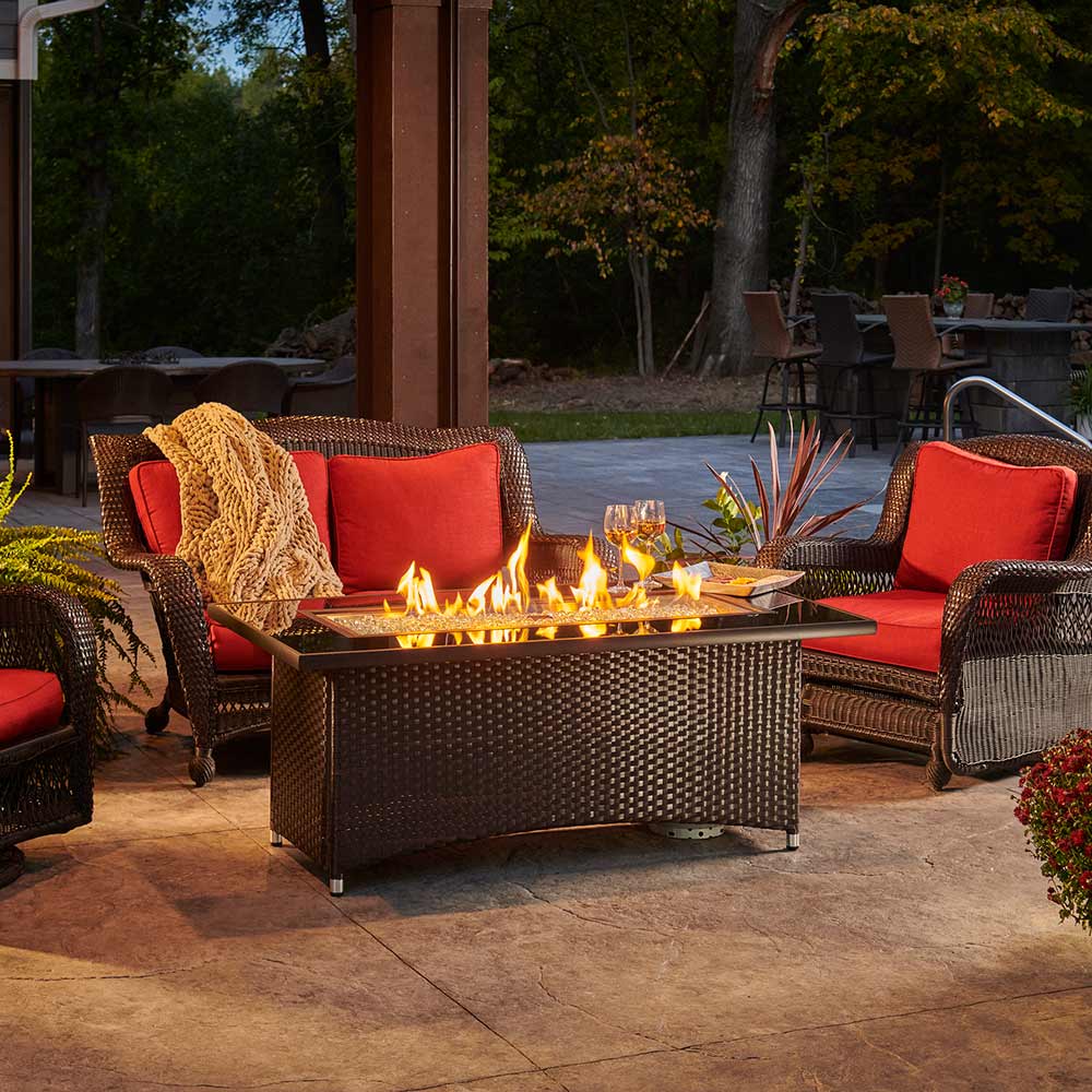 Outdoor gas fire table centerpiece with visible flames, glass windscreen, and dark woven body