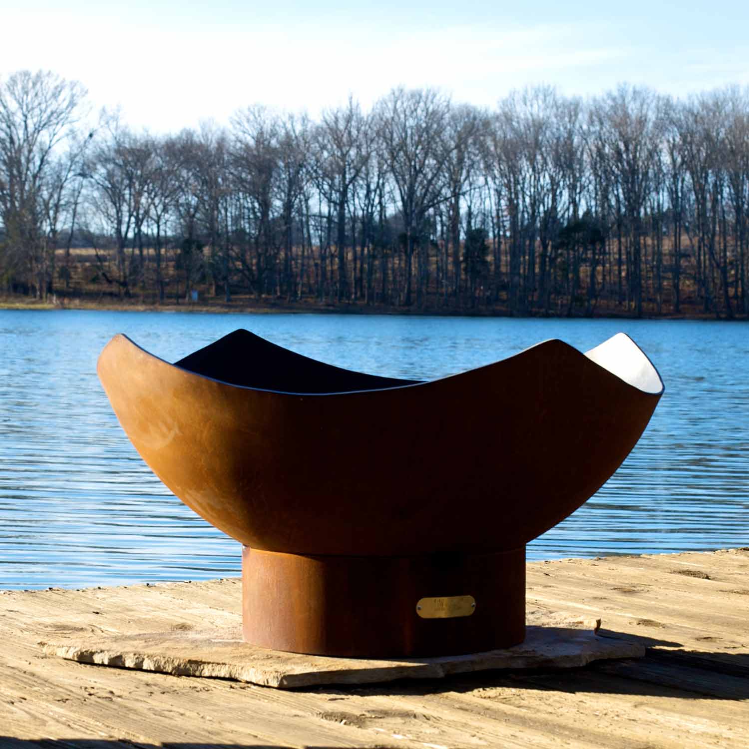 Manta ray gas fire pit in bronze with black interior, set by a lakeshore