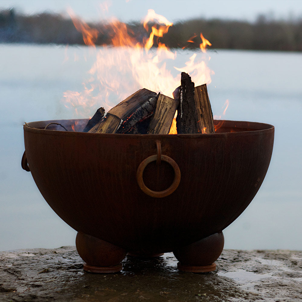 Wood fire pit bowl with flaming logs and ring handle, dark copper-brown metal.