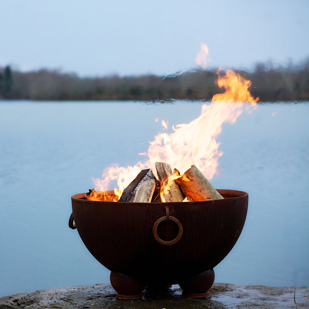 Wood fire pit with burning logs and ring handle, standing on stone by a lake.