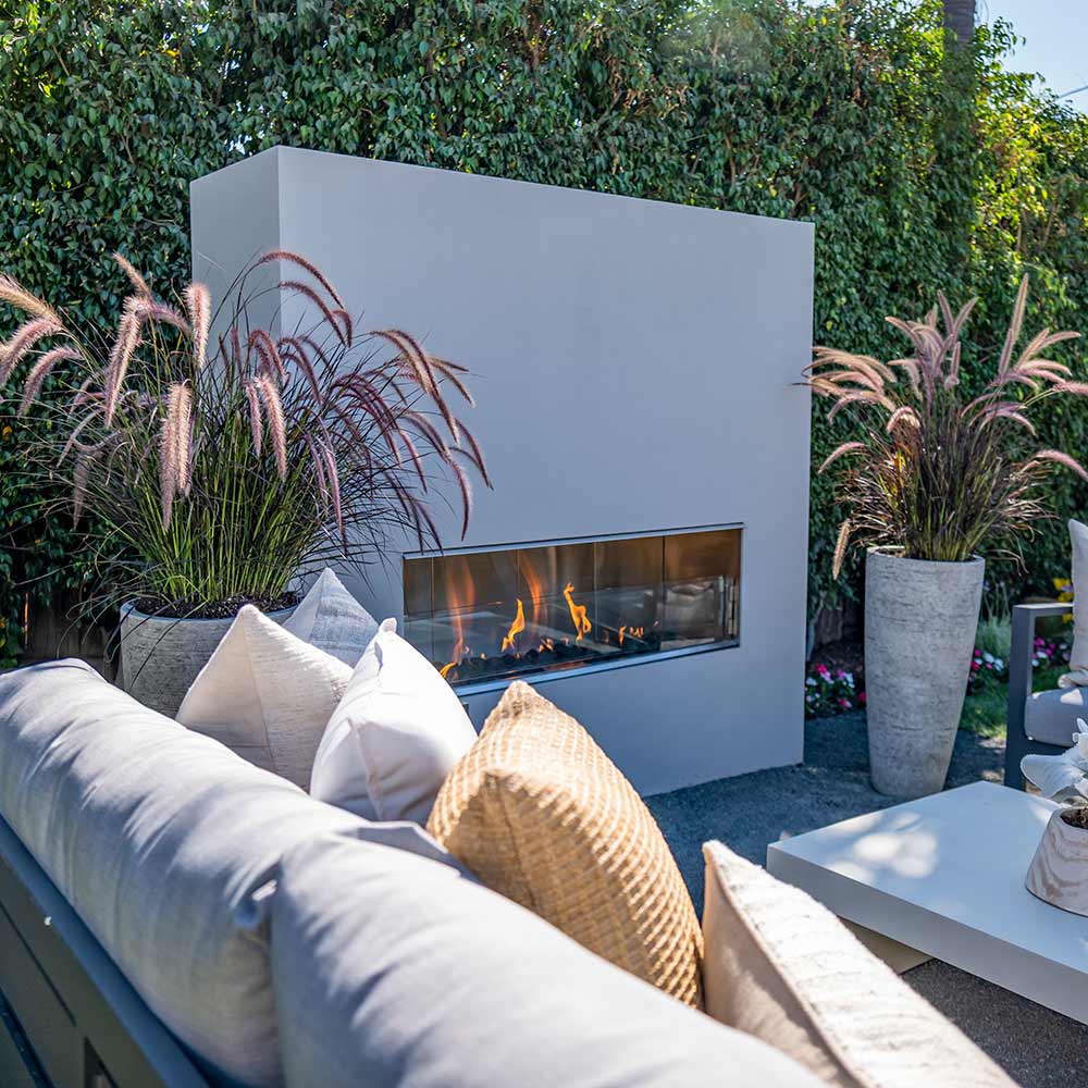 Outdoor linear fireplace set into a light wall with glass front and flames.