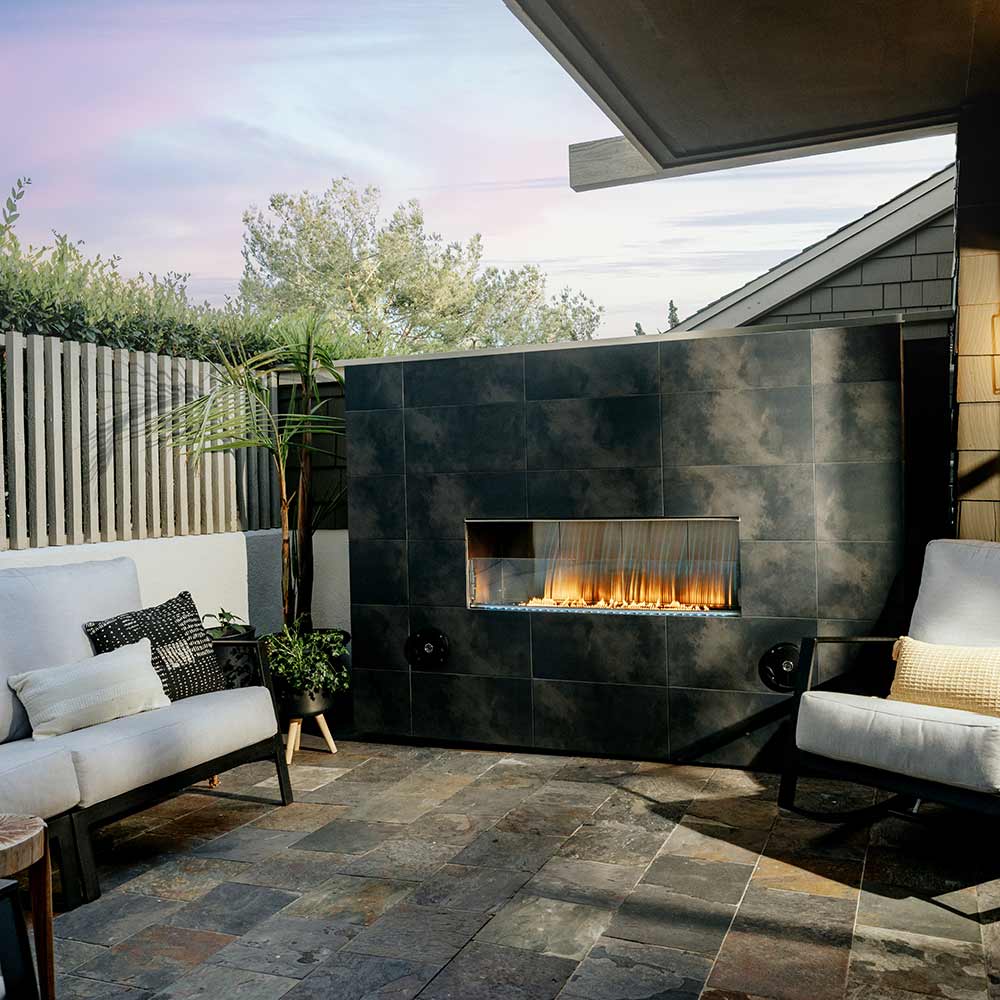Outdoor fireplace set into stone wall, glass window showing flames, dark metal body