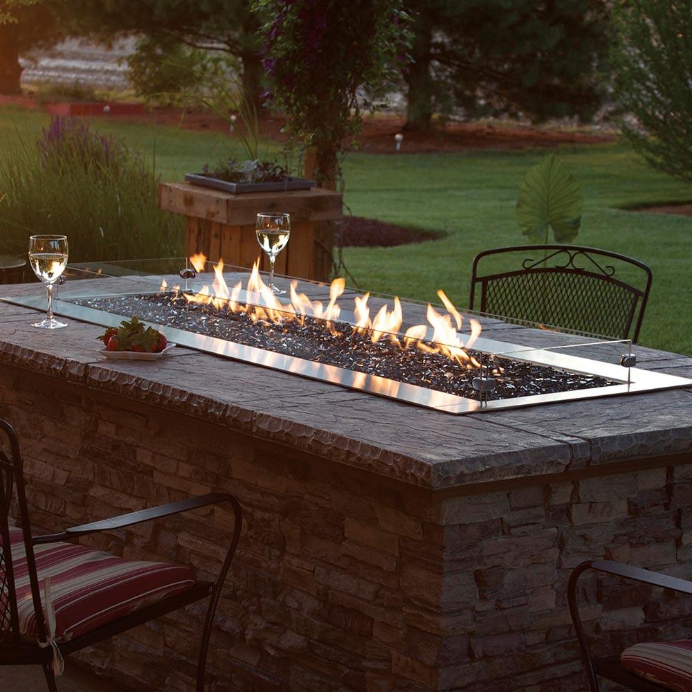 Outdoor linear gas fire pit with flames and dark burner media on stone surround