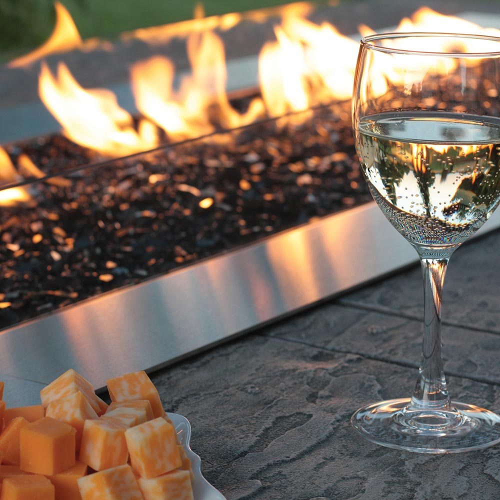 Outdoor linear gas fire pit flames over dark burner tray with wine glass nearby
