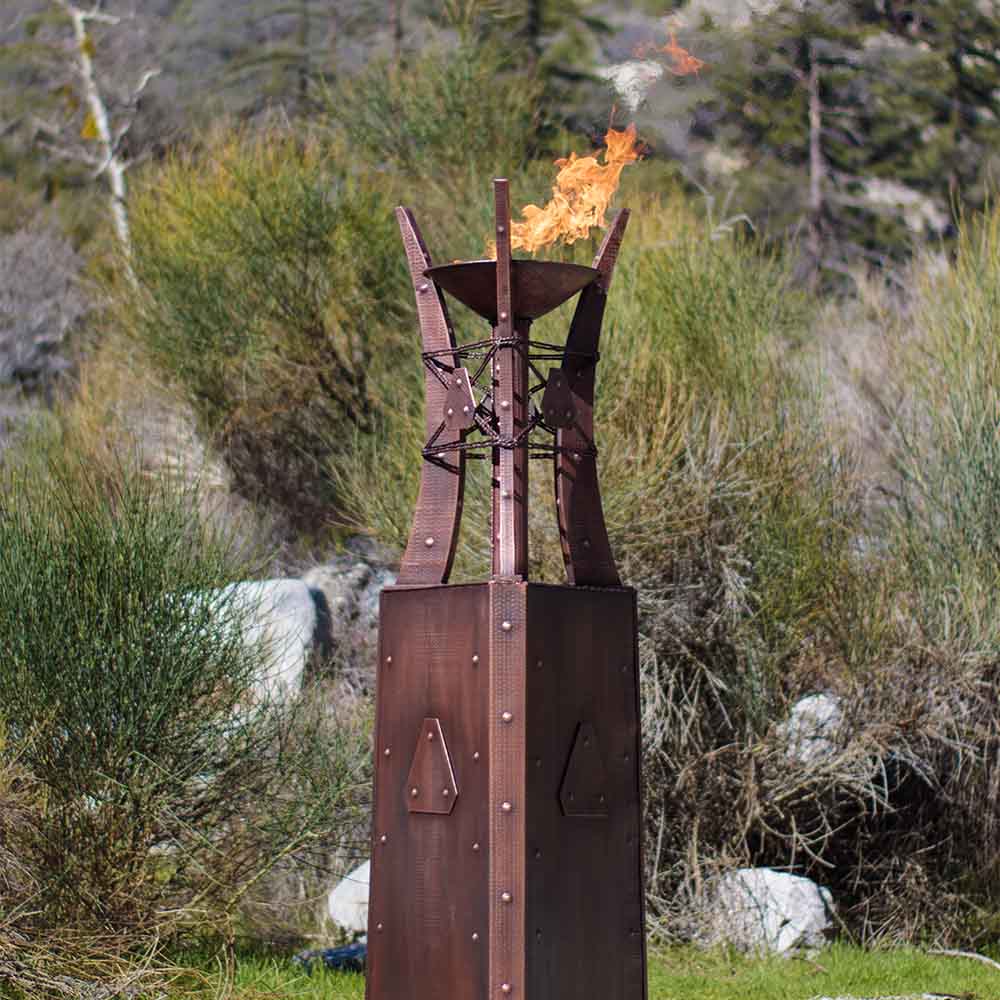 Copper fire tower with visible flames outdoors near shrubs and rocks