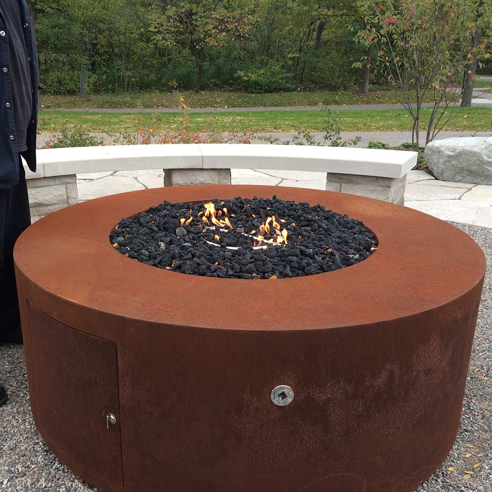 Round corten steel gas fire pit with flame in a black lava rock bowl