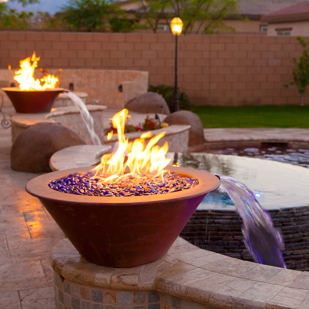 Round gas fire bowl on outdoor stone surface with flame and water feature beside it
