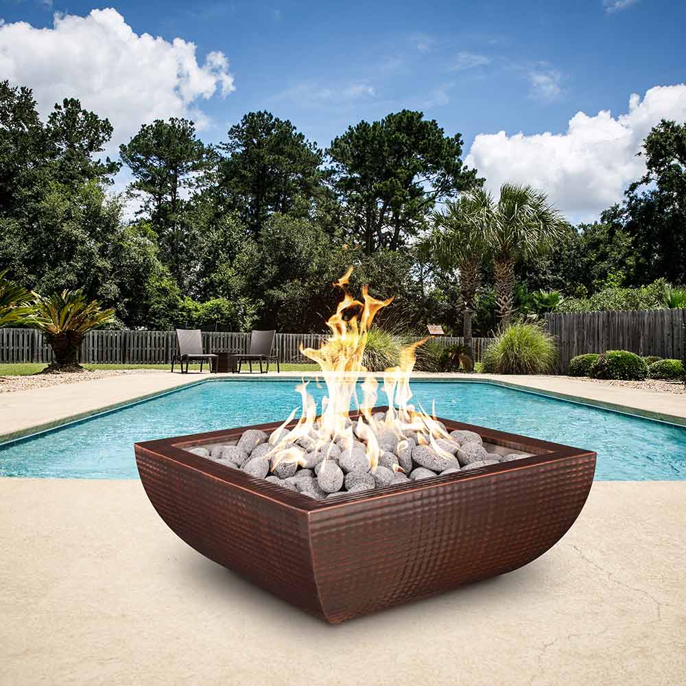 Copper gas fire bowl with flames over gray stones beside a pool