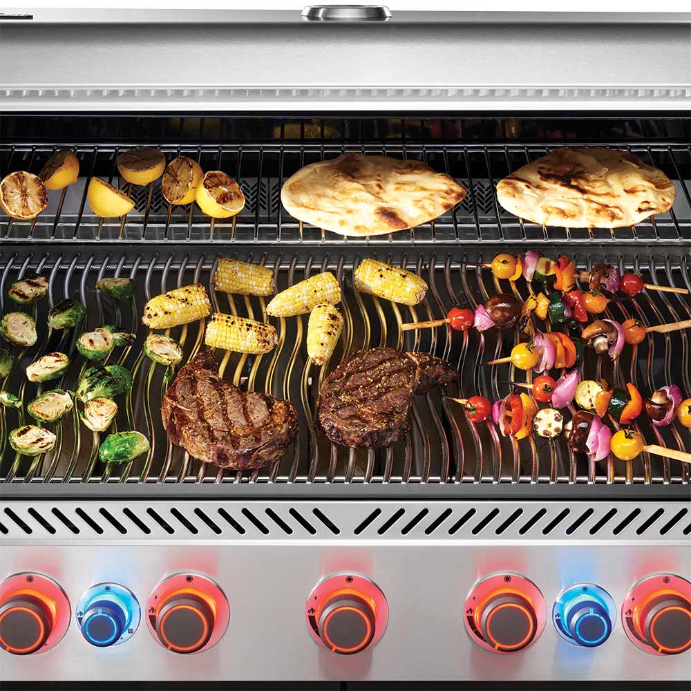 Grill grates with steaks and vegetables cooking over gas burners.