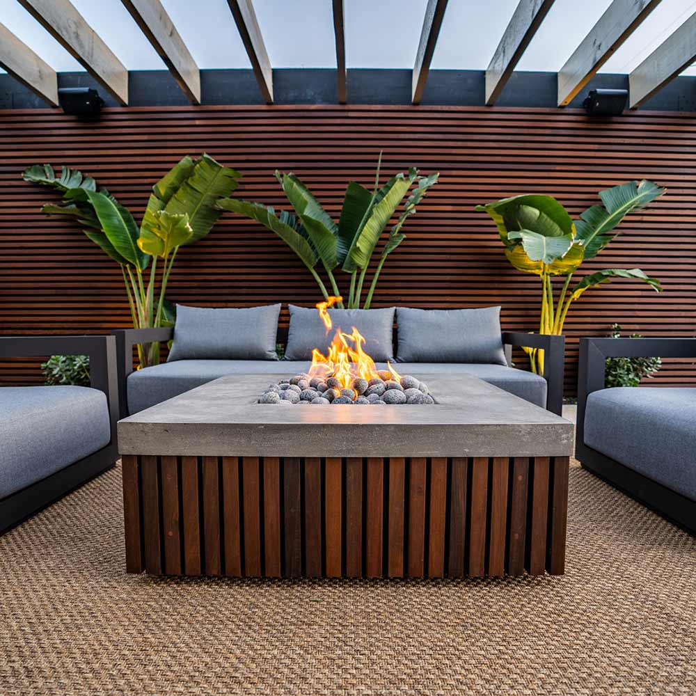 Gas fire pit with concrete top, wooden slat base, and visible flames in the center.