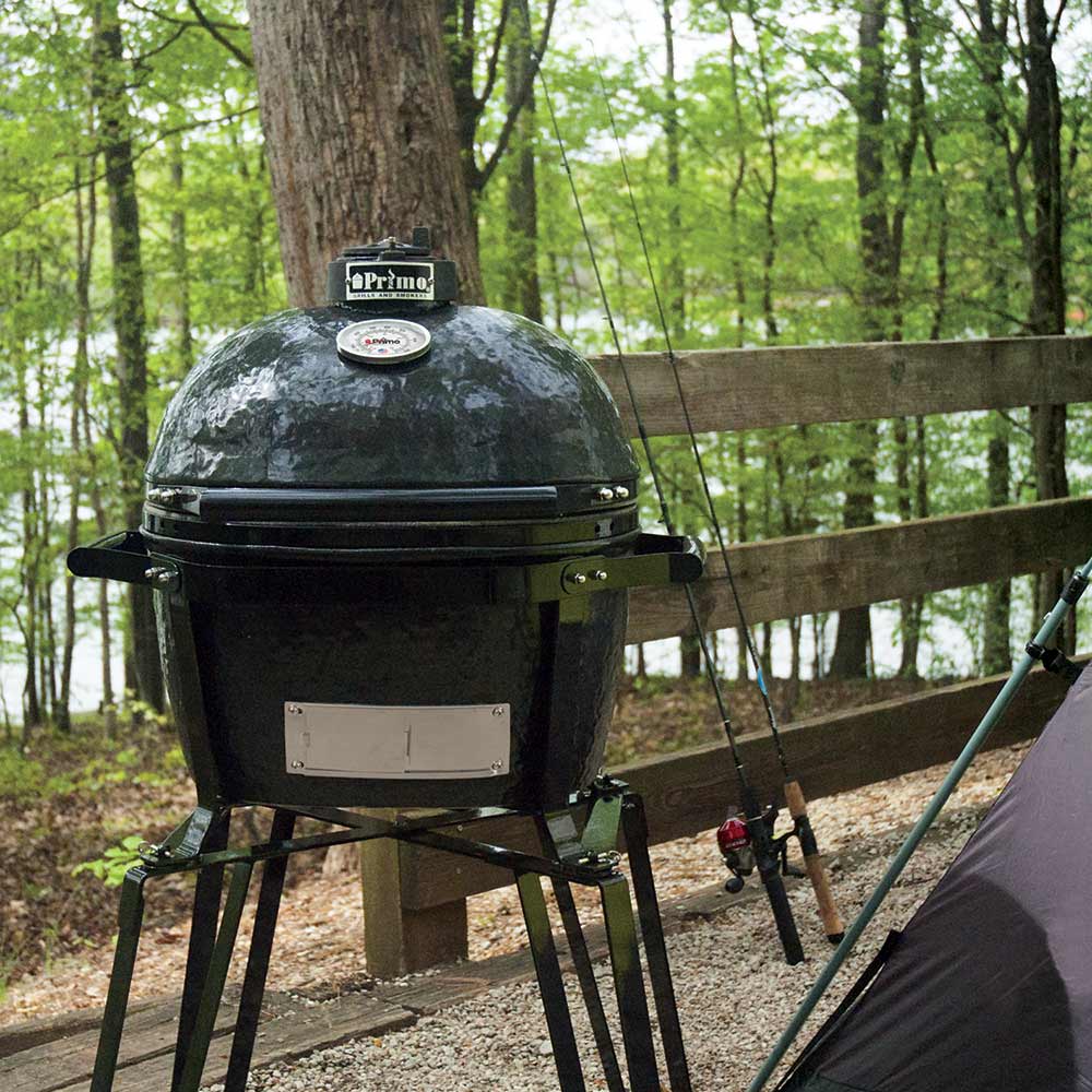 Black Primo grill on a stand outdoors with trees and a lake in the background