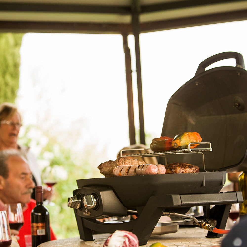 Portable tabletop gas grill on stand, lid open with food cooking on the grate.