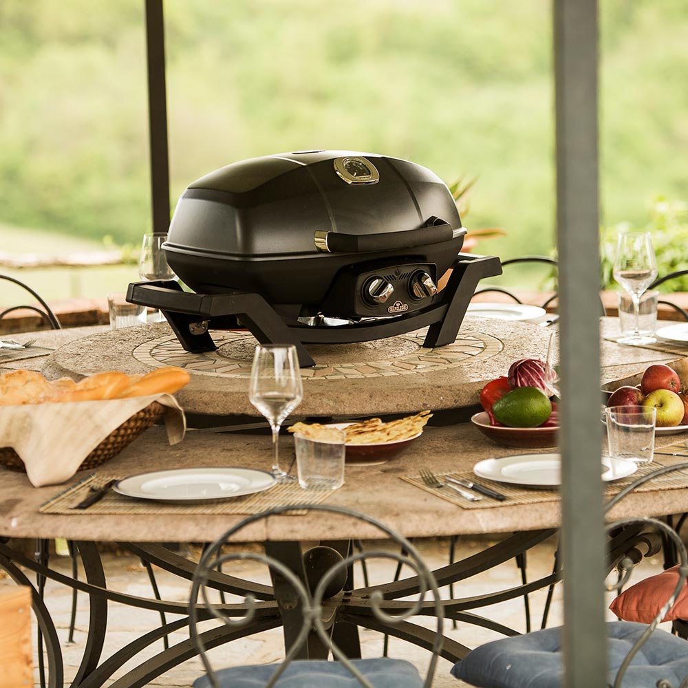 Portable tabletop gas grill on outdoor table, lid closed, with side controls visible.