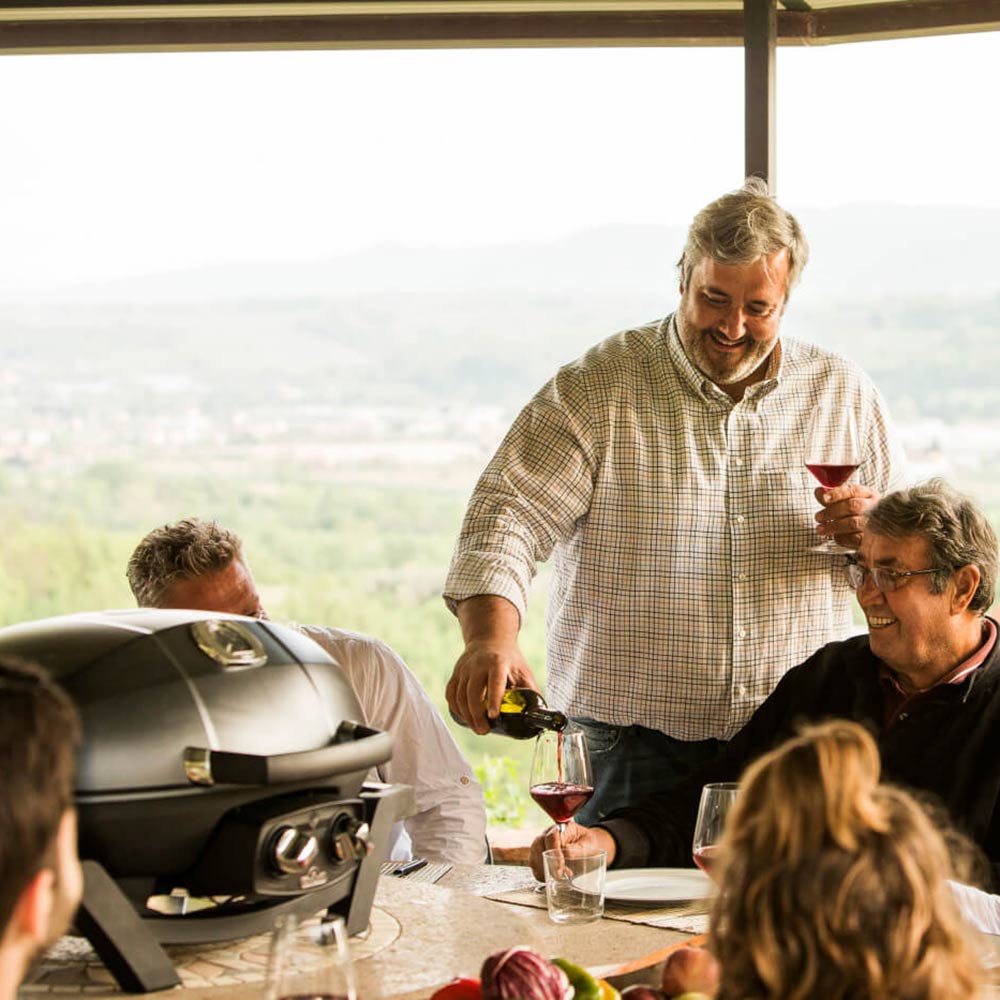 Portable tabletop gas grill on outdoor table with people seated nearby, lid open.