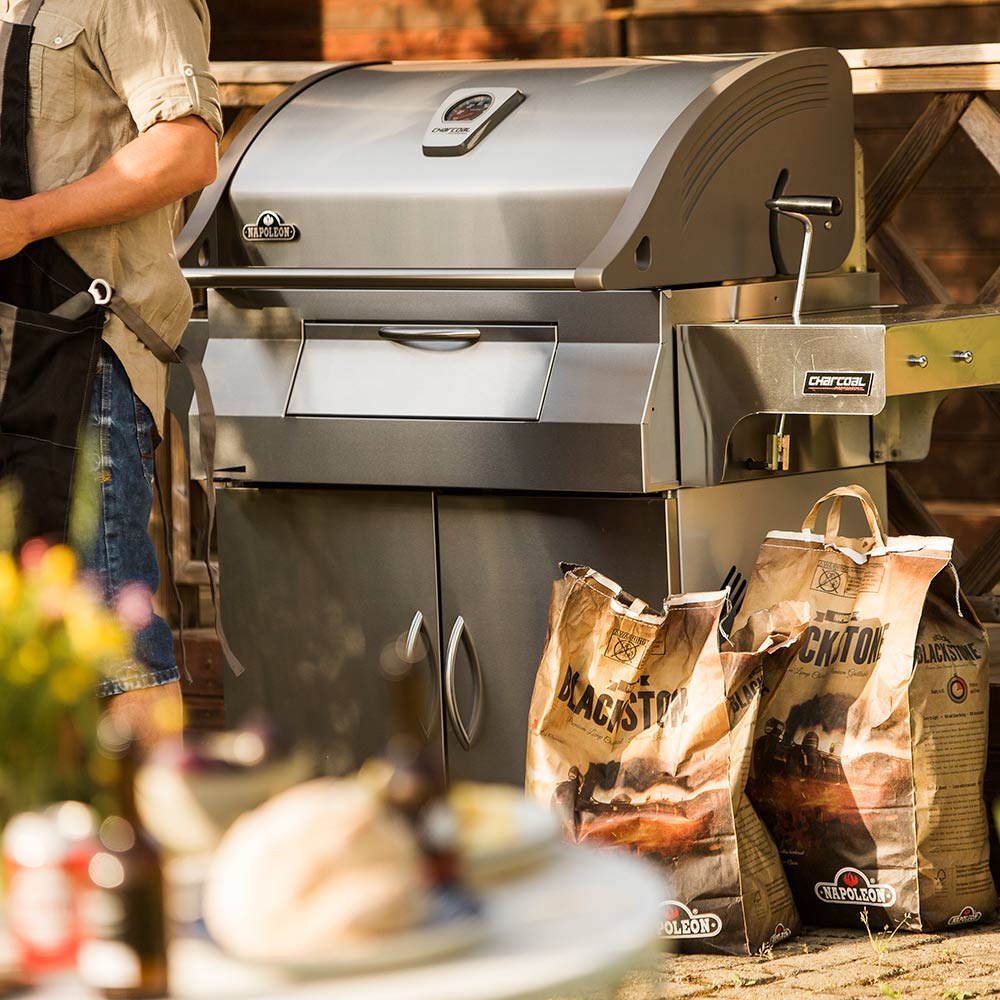 Charcoal grill on cart with domed lid and fuel bags on the side shelf