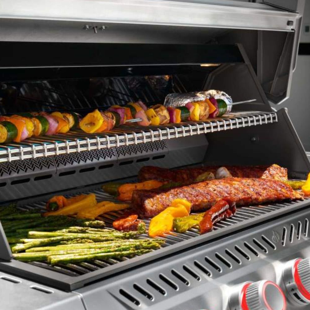 Open gas grill with seasoned food on grates and a metal warming rack