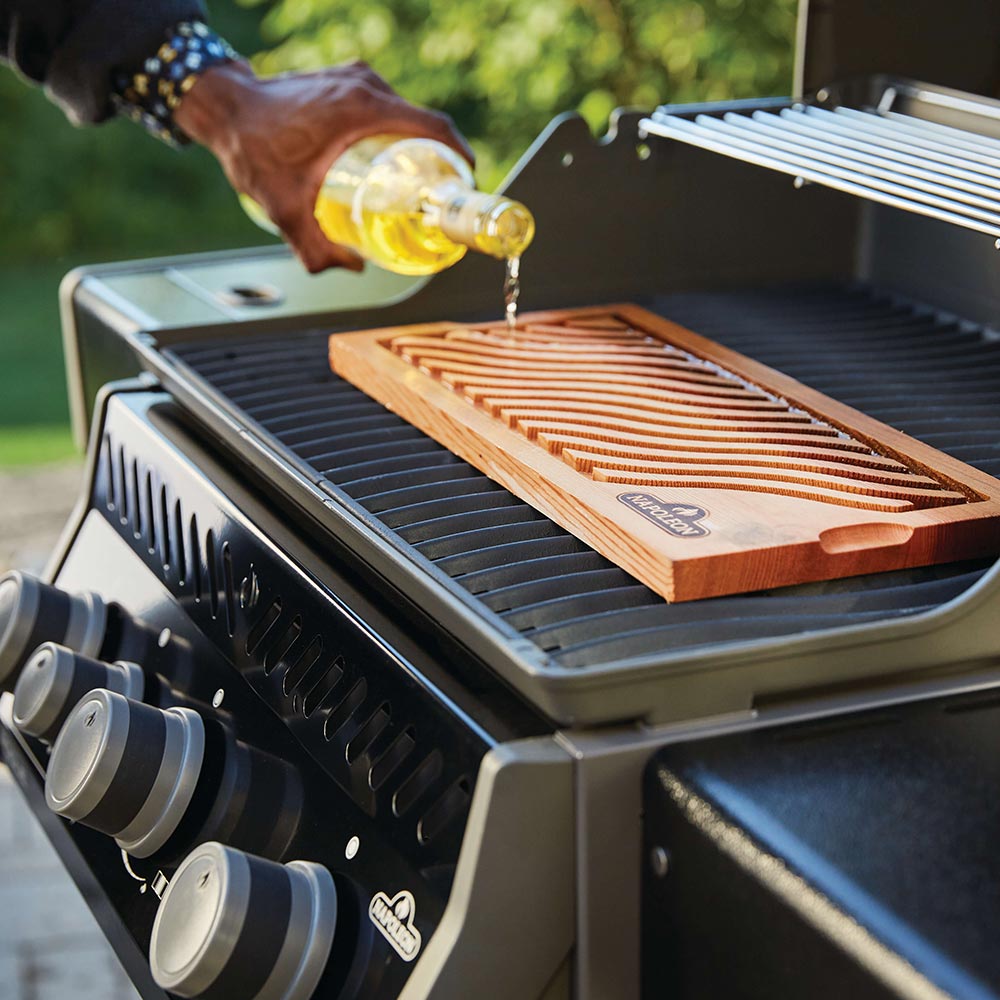 Gas grill grates with a wooden plank and oil being poured onto food
