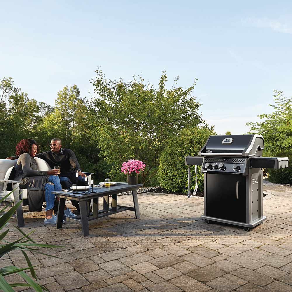 Black gas grill on cart standing outdoors with lid closed and side shelves
