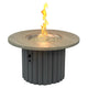 Round gas fire pit table with light wood top and dark fluted base; flames rising from bowl.