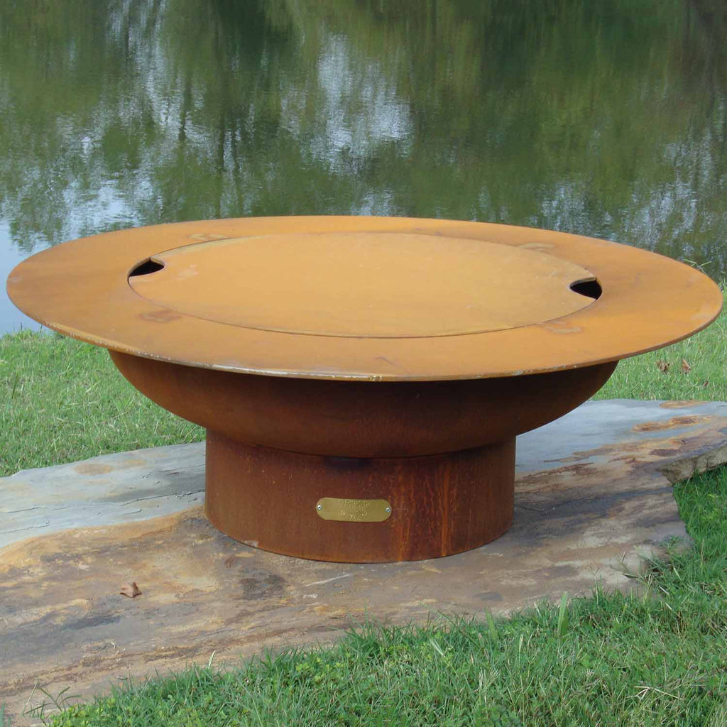 Round gas fire pit with closed lid, tan outer body and brass nameplate on stone surface