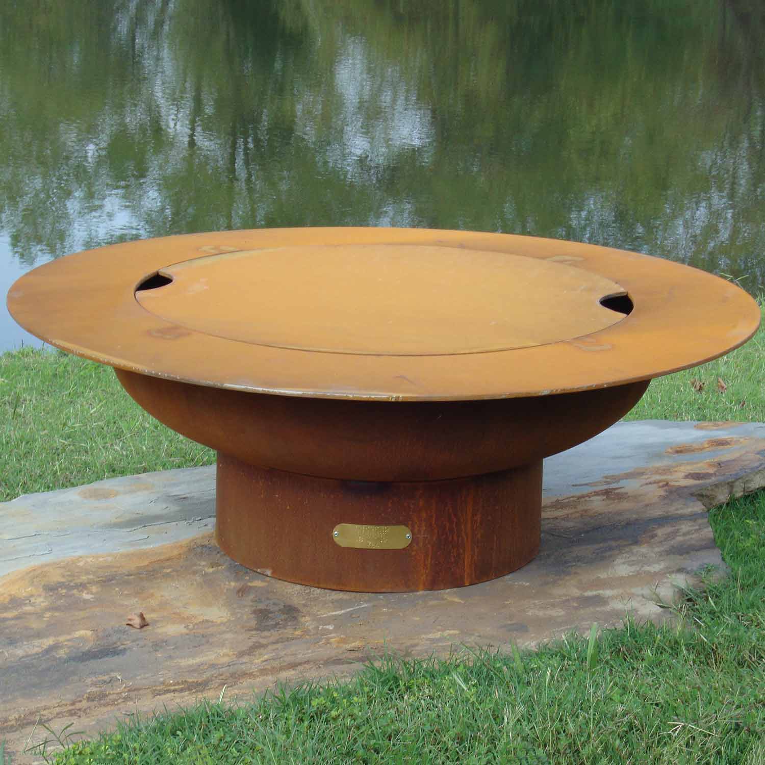Round gas fire pit with closed lid, tan outer body and brass nameplate on stone surface