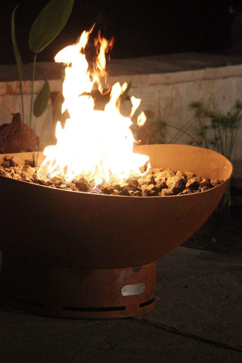 Rust-orange gas fire pit with tall flames and rock-filled burner basin.