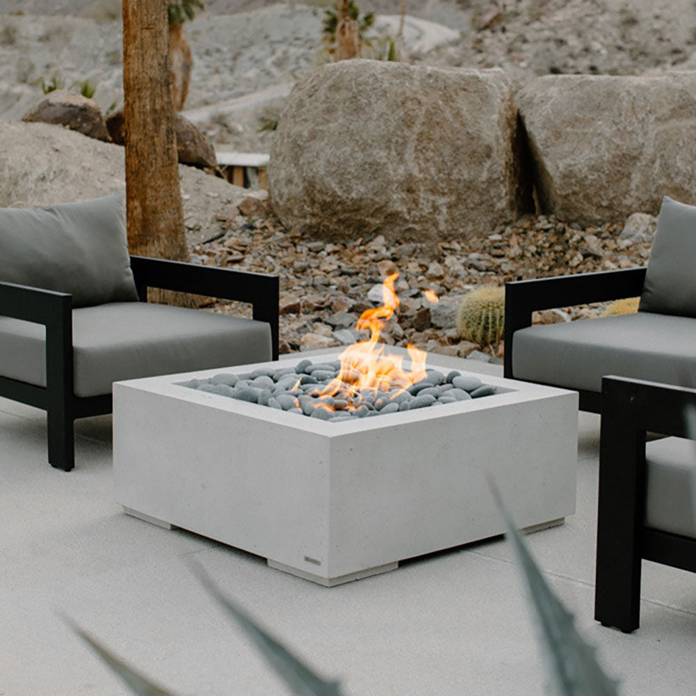 Square concrete gas fire pit with pebble stone bed and small flames in the center