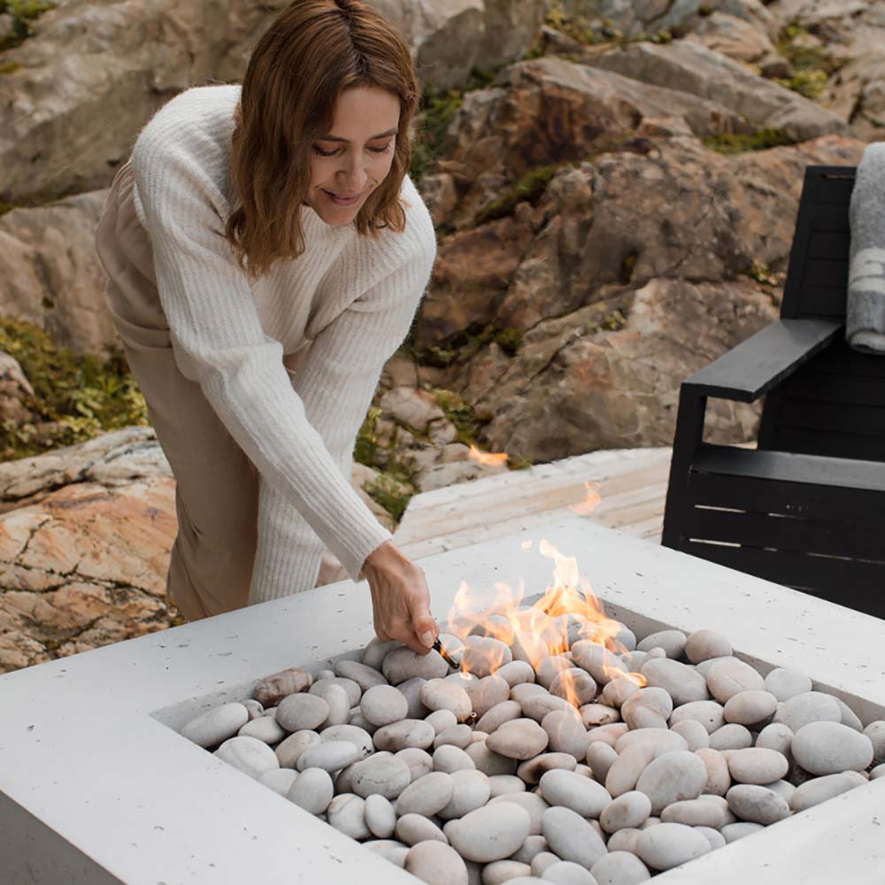 Square concrete gas fire pit with flames and stones, hand adjusting fuel