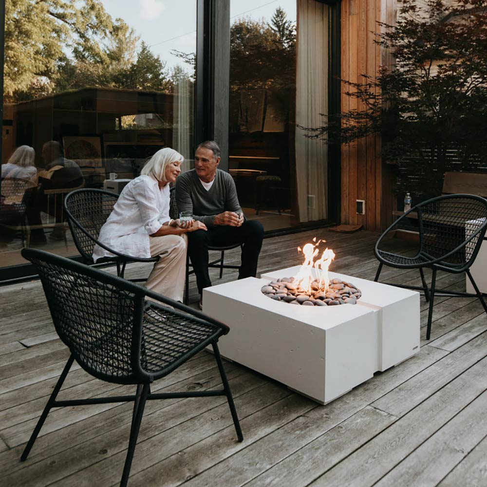 Square concrete gas fire pit with flame and rocks on an outdoor patio near seating