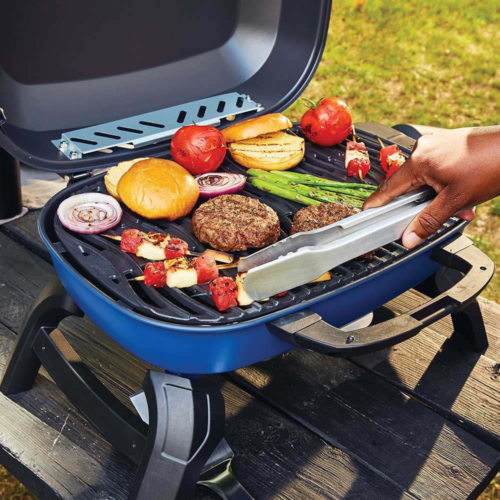 Open blue portable gas grill cooking grate with burgers, vegetables, and onions