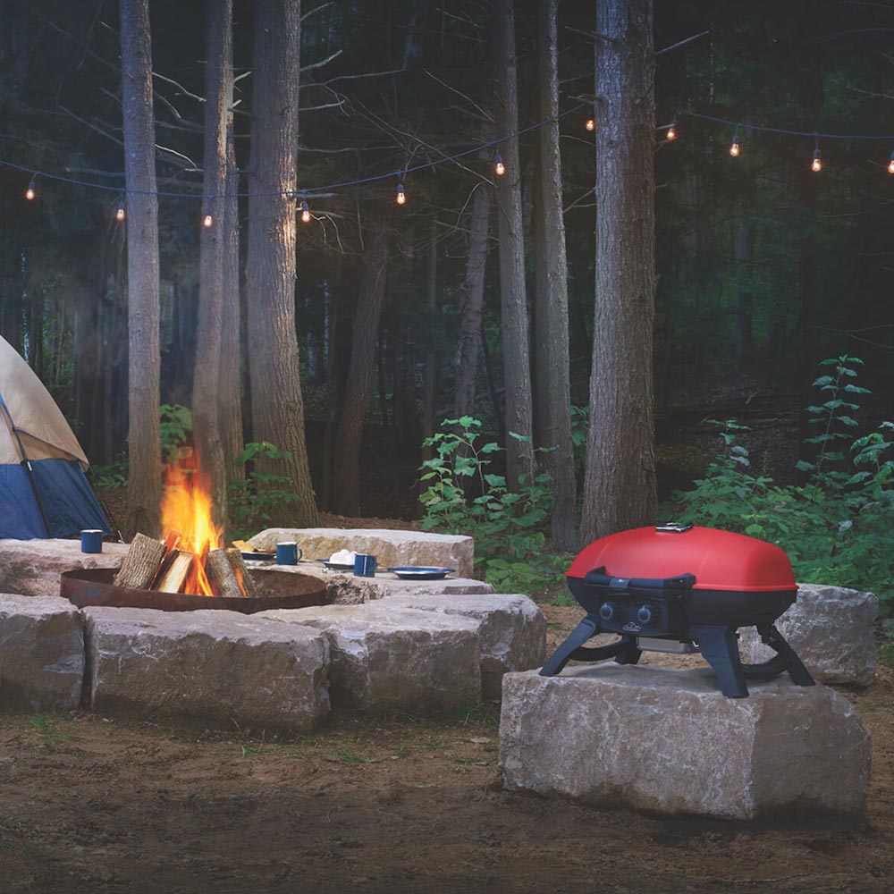 Red portable tabletop gas grill on stone blocks near a campfire in the yard