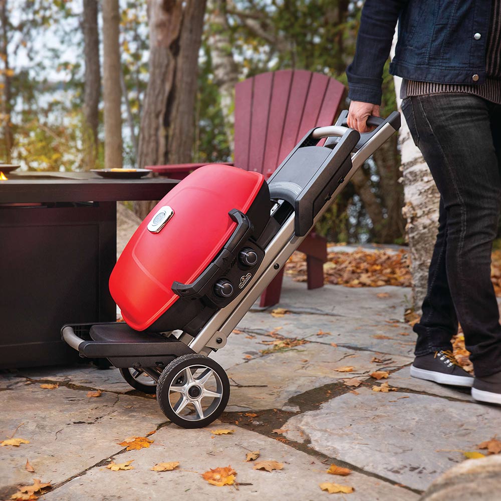 Red portable propane grill being pushed on its cart with wheels outdoors.