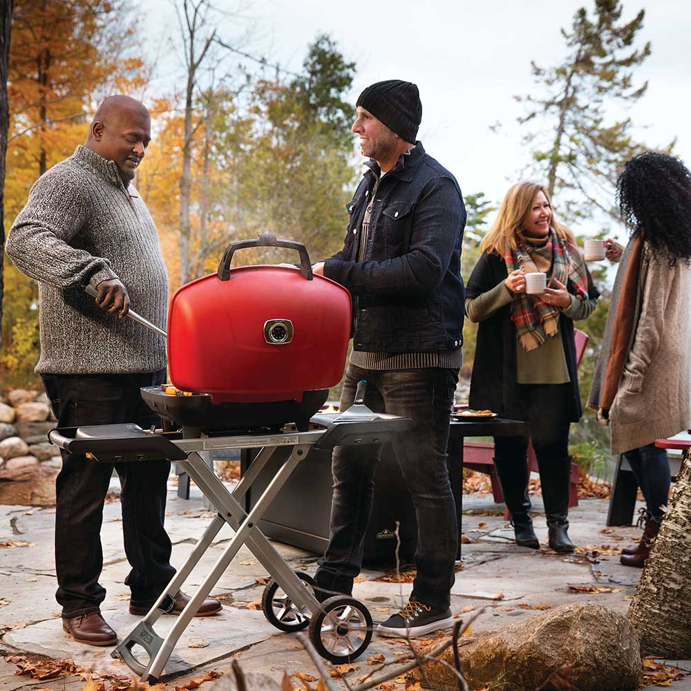 Red portable propane grill on folding cart outdoors with people gathered nearby.