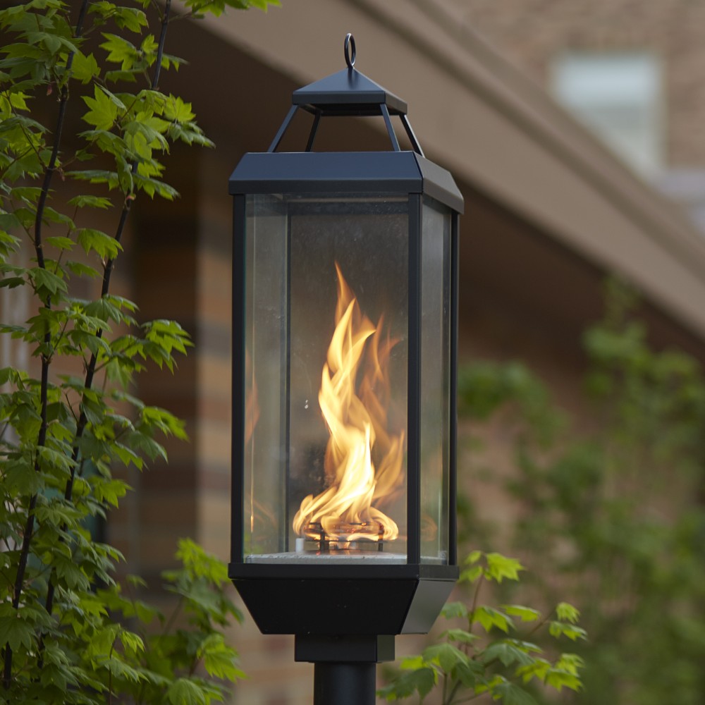 Black gas lantern on outdoor post mount with visible flame inside glass