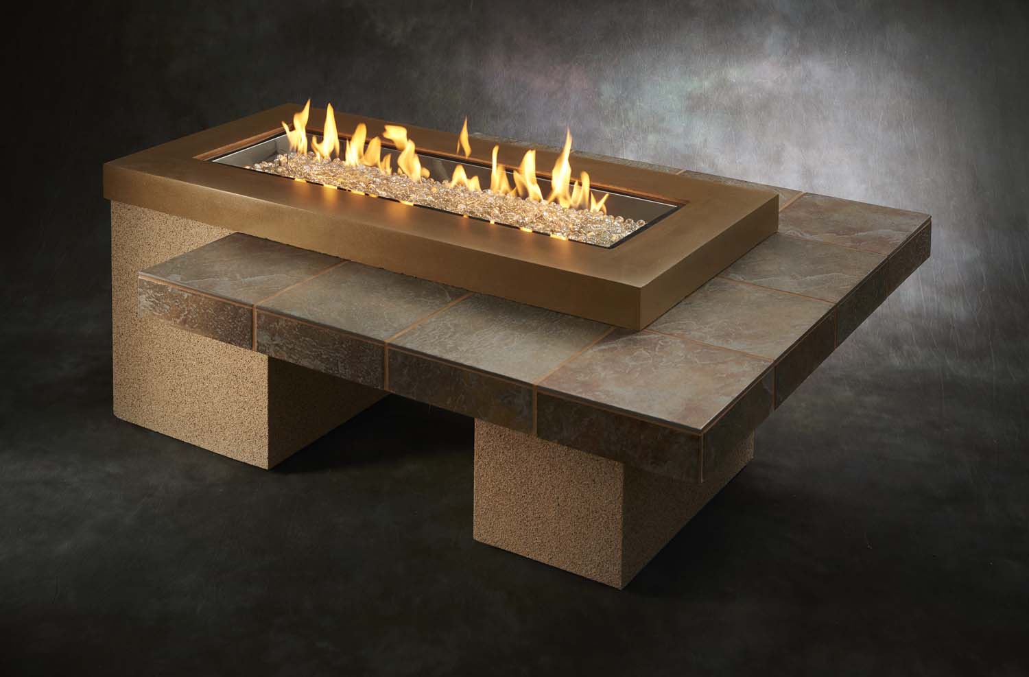 Linear gas fire pit table with tan stone top and matching stone-look base; flames visible.