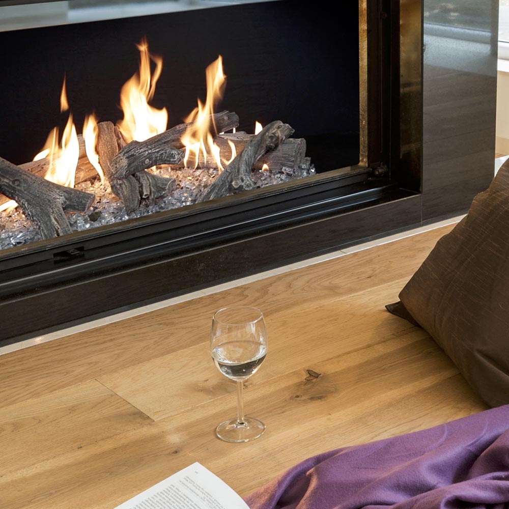 Linear gas fireplace with burning dark logs behind glass and hearth media stones