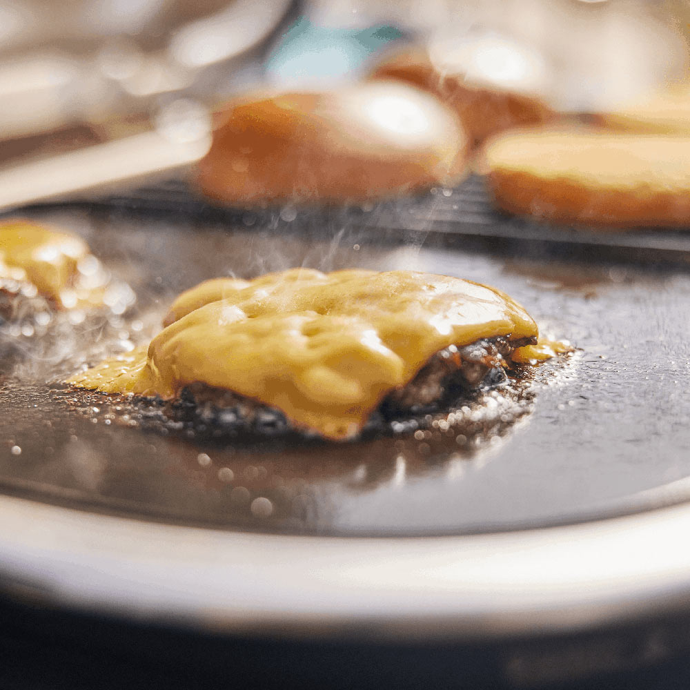 Griddle cooking surface with sizzling patties and sliced bread, rising steam