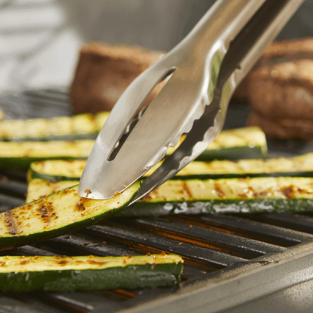 Grill tongs holding zucchini slices on a hot grill grate