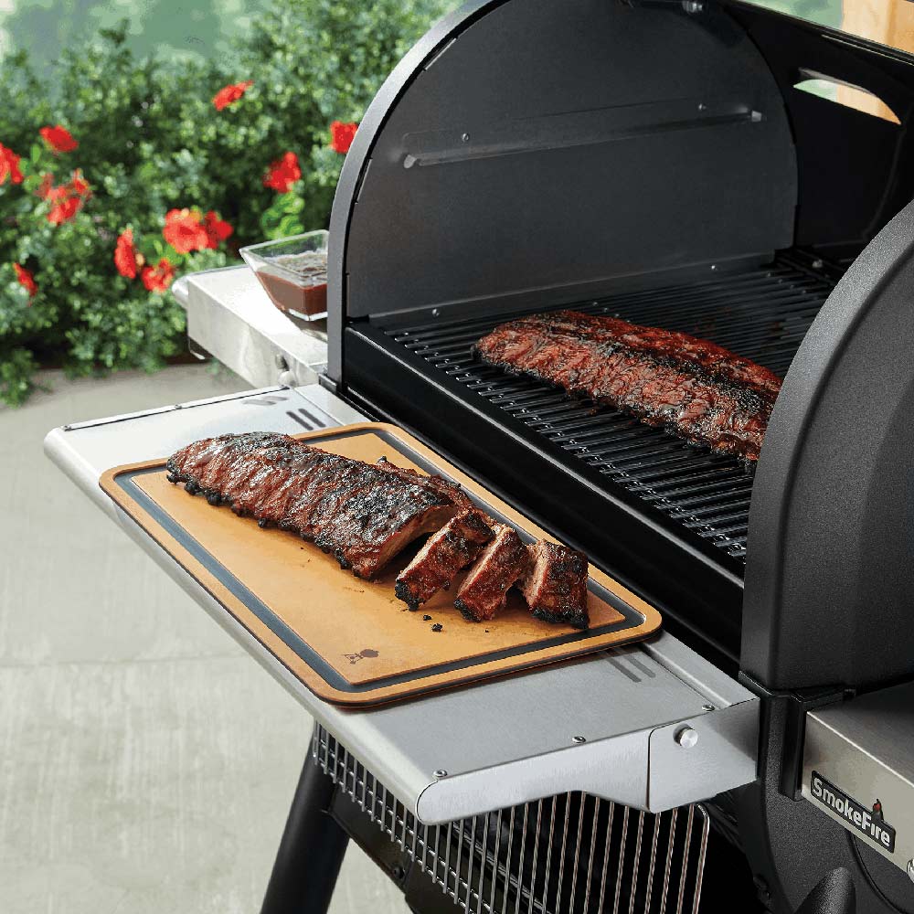 Cutting board with tan surface and dark border, resting on a grill ledge with sliced meat.